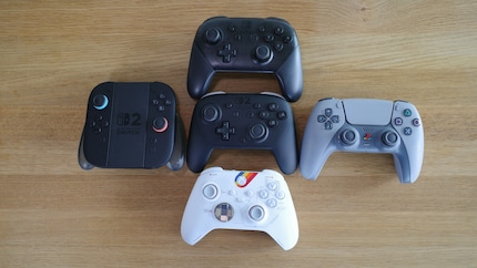 Der Switch 2 Pro Controller (mitte) im Vergleich zu: Switch 1 Pro Controller (oben), PS5-Controller (rechts), Xbox-Controller (unten), Joy-Con-2-Controller in Aufladehalterung (links).
